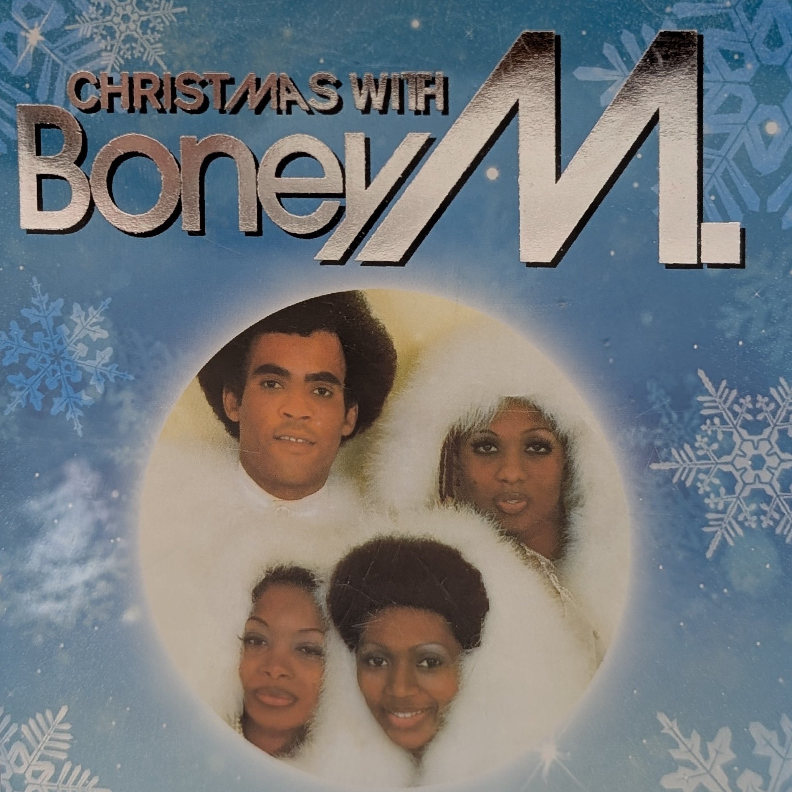CD cover for 'Christmas with Boney M.' featuring the group's name in large metallic lettering over a blue winter-themed background with snowflakes, and a circular photo of four members in white fur coats.