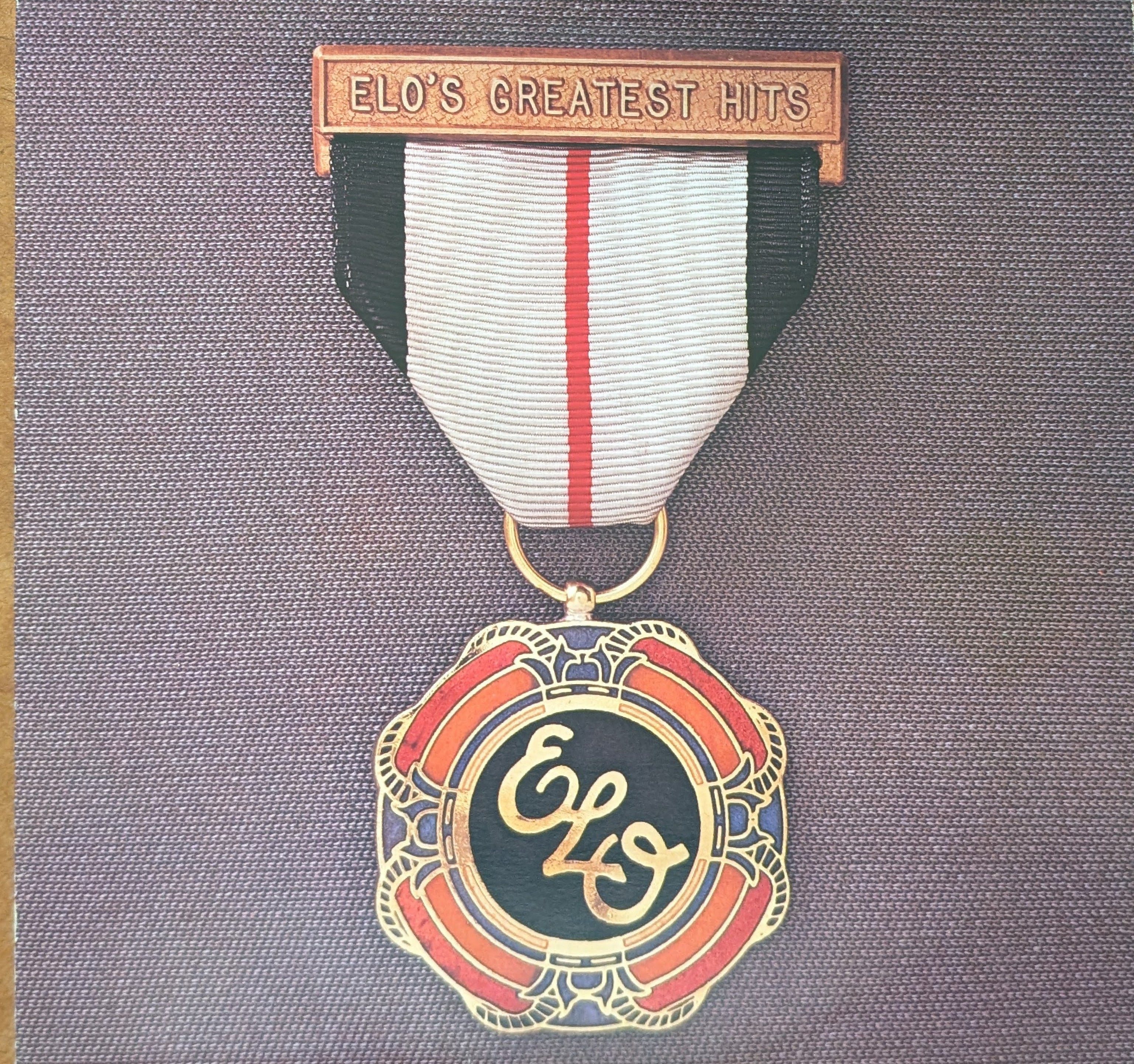 A military-style medal pendant with a circular gold-toned top medallion featuring red, orange, and black enamel, suspended from a ribbon with black, white, and red sections, and a bronze-toned bar at the bottom reading 'ELO'S GREATEST HITS'.