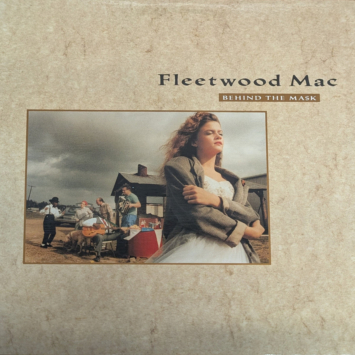 Fleetwood Mac 'Behind the Mask' vinyl record album cover, featuring a textured beige background with a central color photograph of the band members in a rustic outdoor setting.