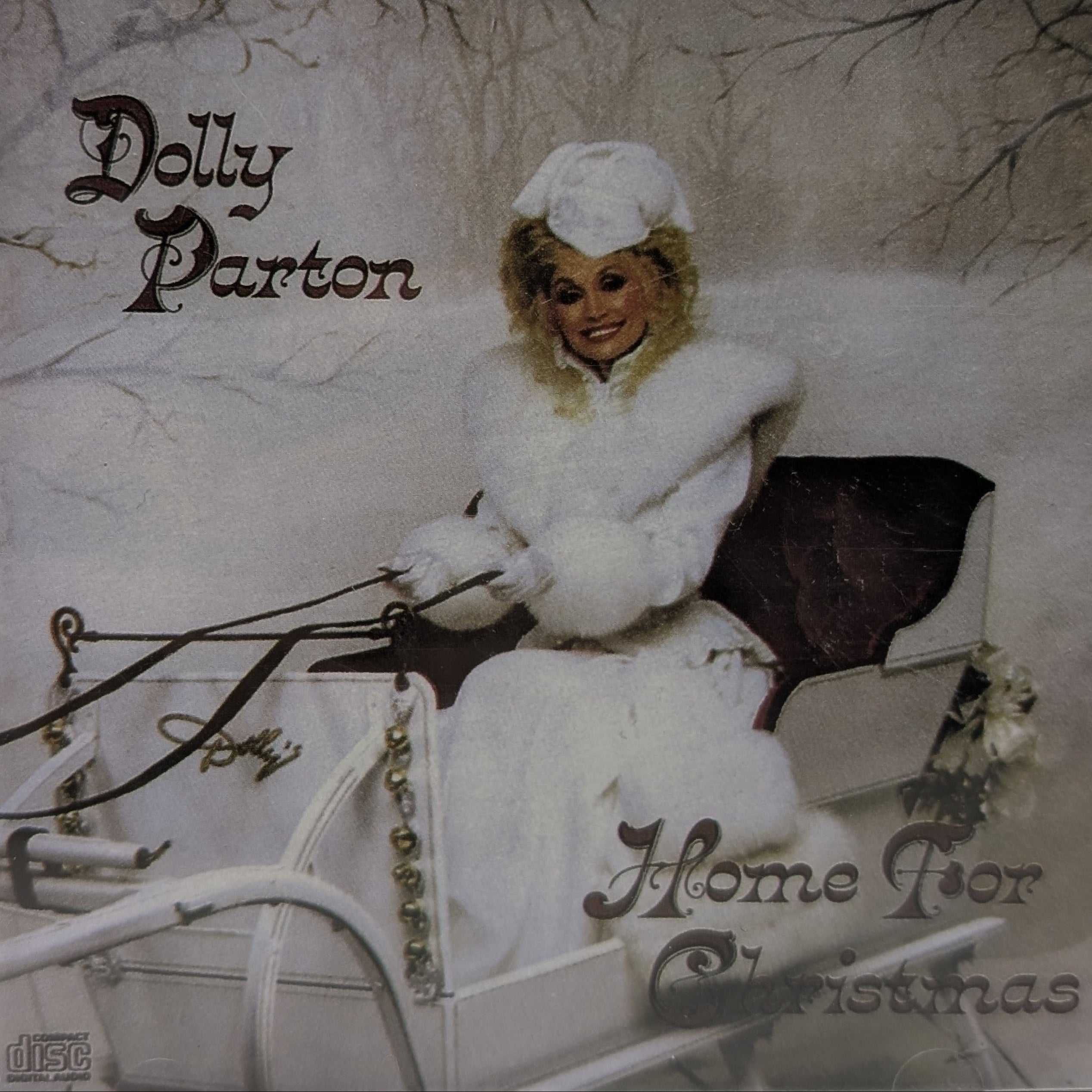 Dolly Parton's 'Home For Christmas' CD album cover, featuring Dolly in a white winter outfit seated in a sleigh against a snowy background.