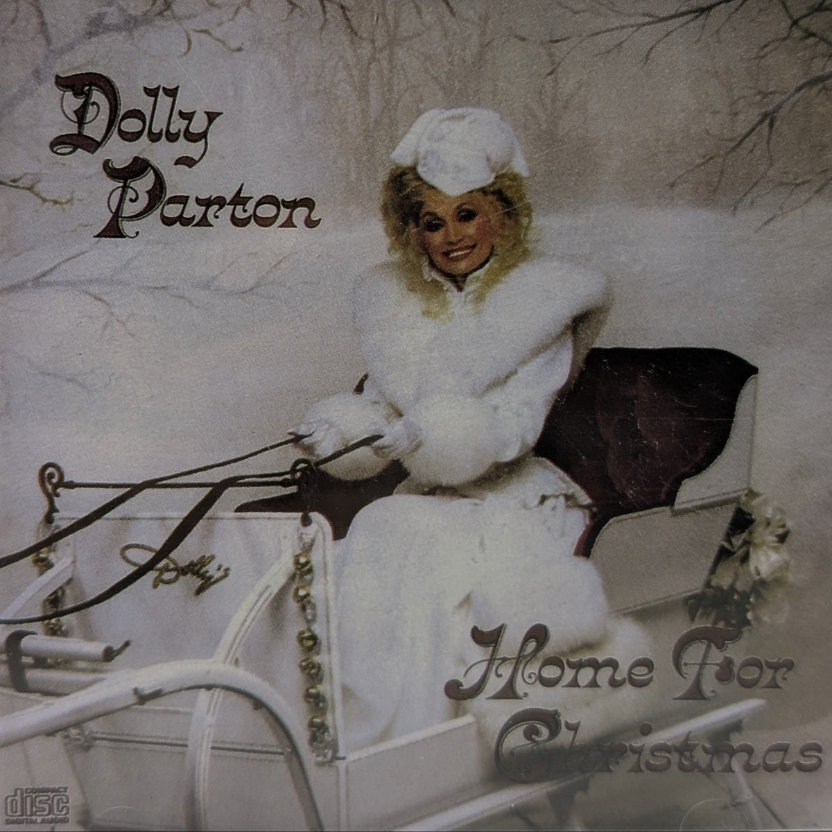 Dolly Parton's 'Home For Christmas' CD album cover, featuring Dolly in a white winter outfit seated in a sleigh against a snowy background.