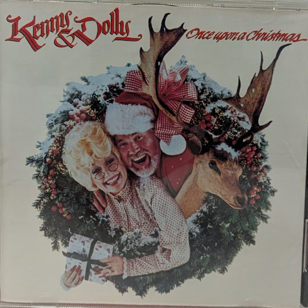 Kenny & Dolly's 'Once Upon a Christmas' CD album cover in a clear jewel case, featuring a festive holiday illustration of a smiling couple in Santa hats surrounded by a snow-dusted wreath and a reindeer.