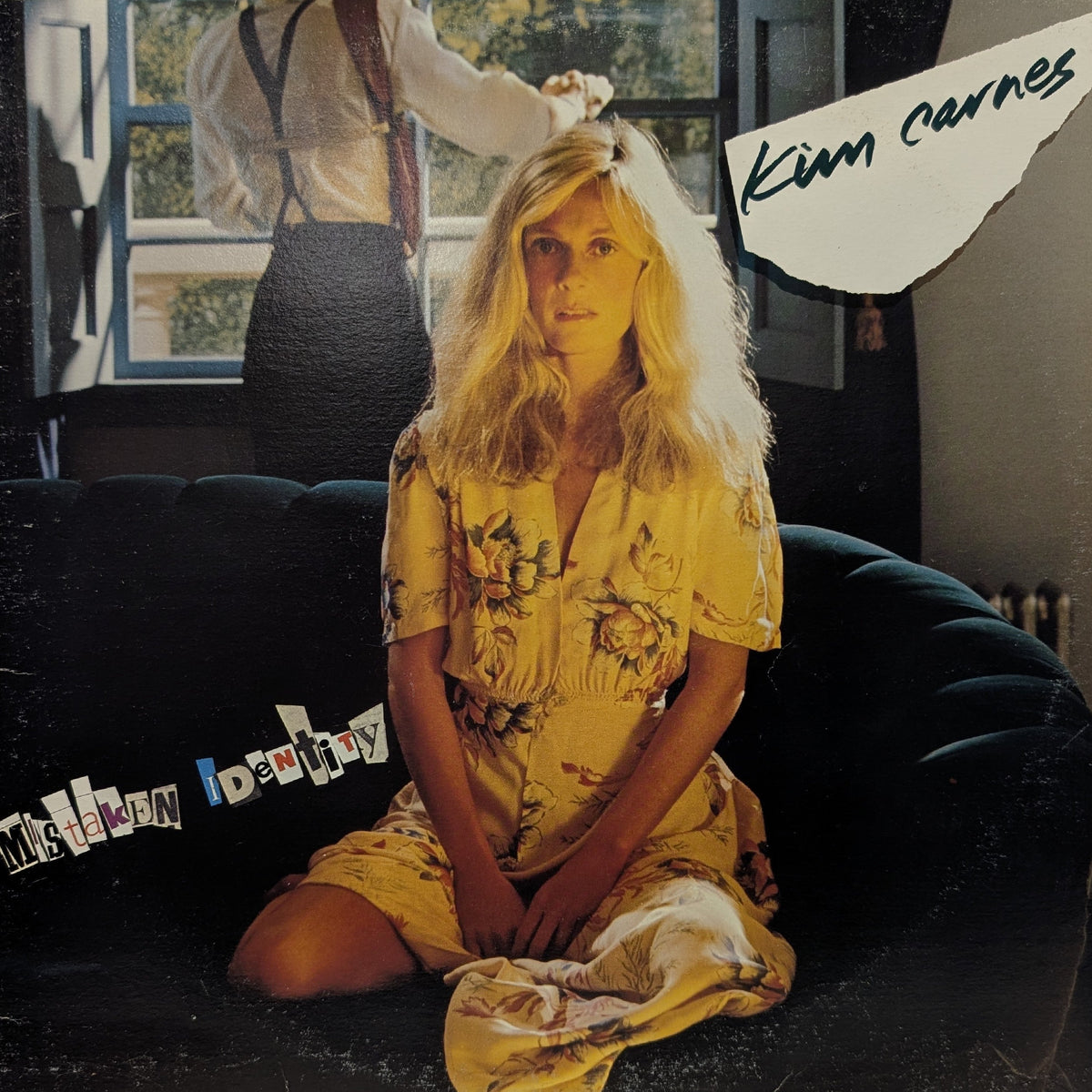 Album cover for Kim Carnes' 'Mishapen Identity', featuring a blonde woman sitting on a dark couch in a yellow floral dress, with a blurred figure in the background.