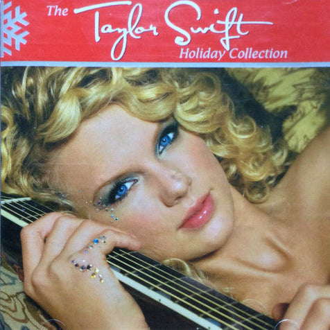 Taylor Swift 'Holiday Collection' CD album in a clear jewel case, resting on a light wood surface. The cover art features a close-up portrait of Taylor Swift with blonde curly hair, blue eyes, and glitter, holding a guitar, with a red header band displaying the album title.